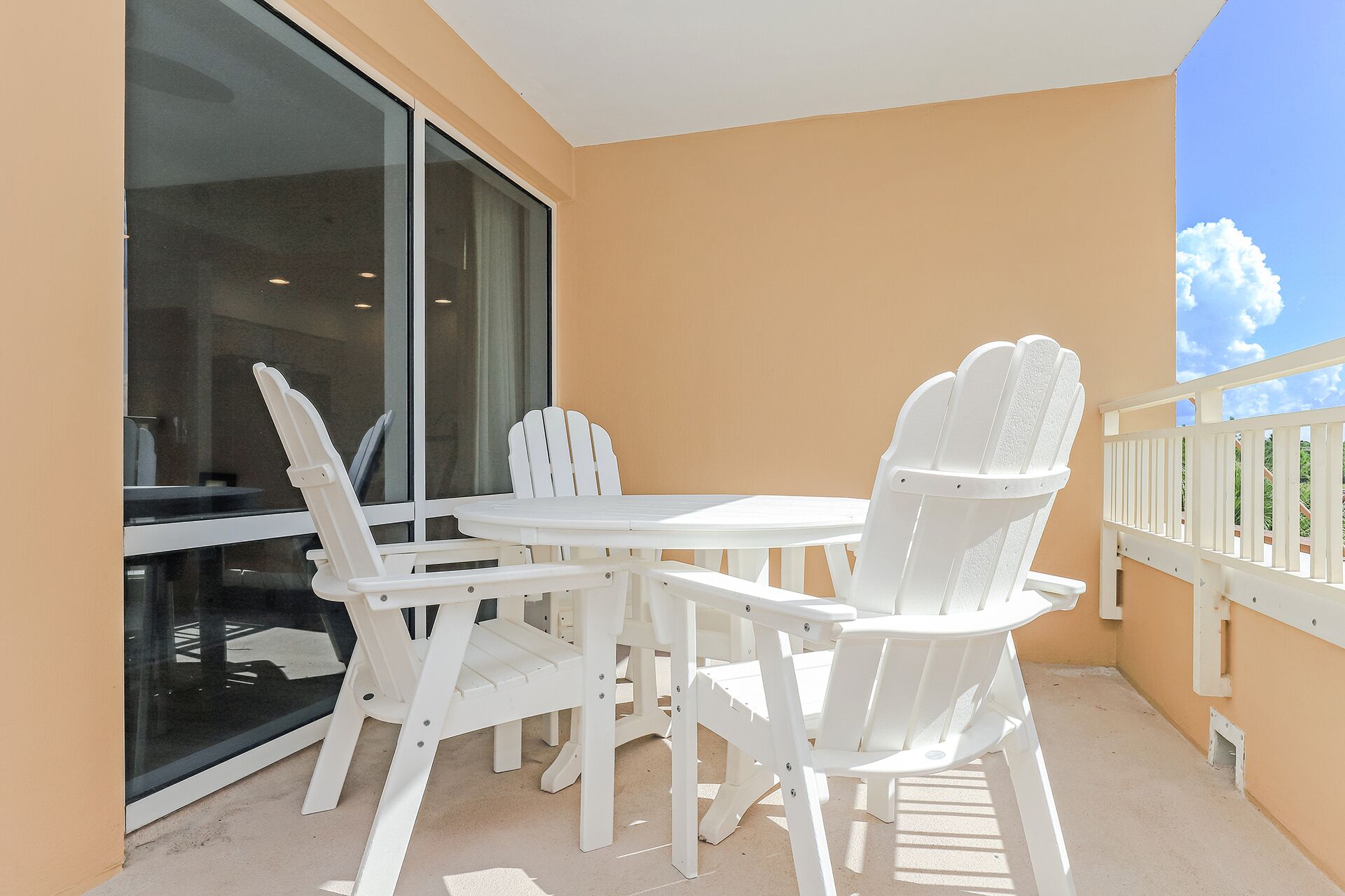 A balcony with white plastic chairs and a table, a glass door, and a view of the sky.