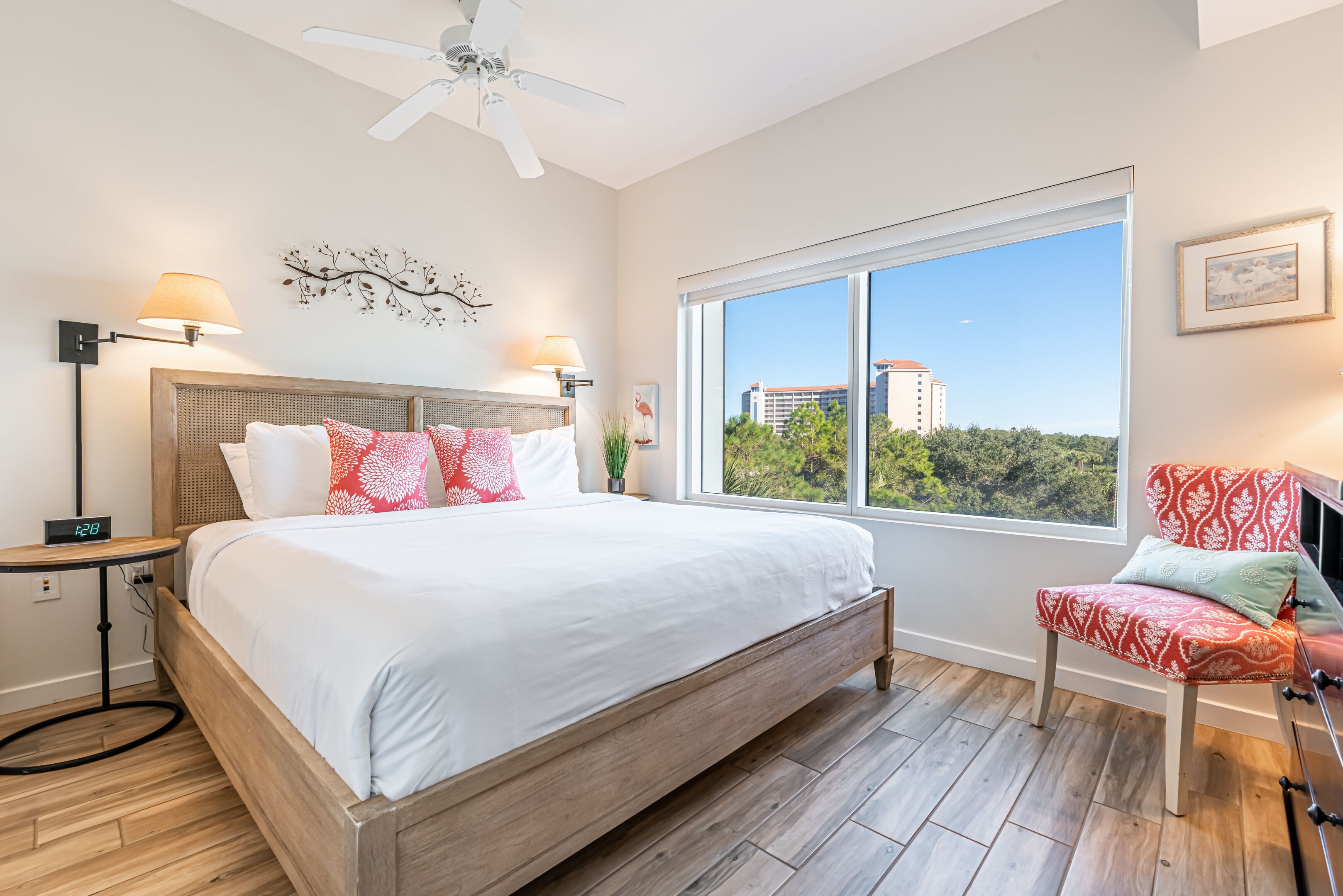Une chambre avec un grand lit, une table de chevet, une chaise, un ventilateur de plafond et une fenêtre donnant sur des bâtiments et des arbres.