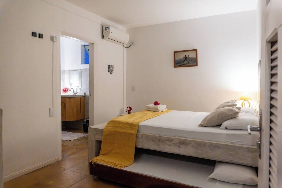 Quarto com cama, mesa de cabeceira com abajur, janela e banheiro ao fundo.