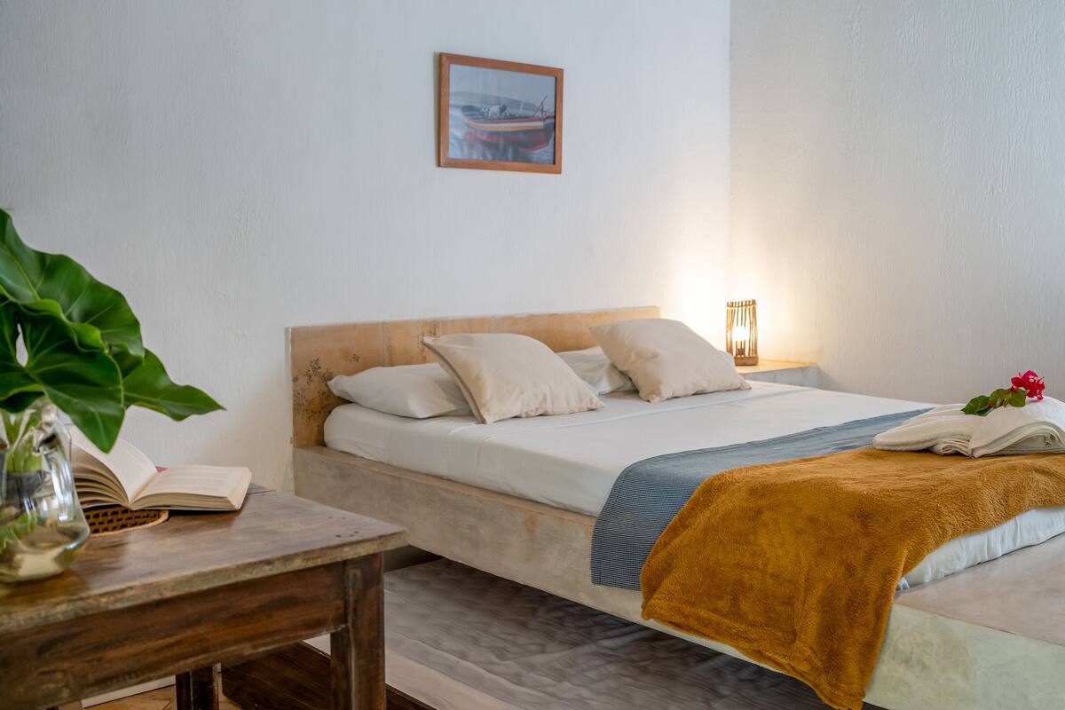 Cama bem arrumada com cabeceira de madeira, mesa de cabeceira com um livro e uma planta, e luminária fixada na parede.