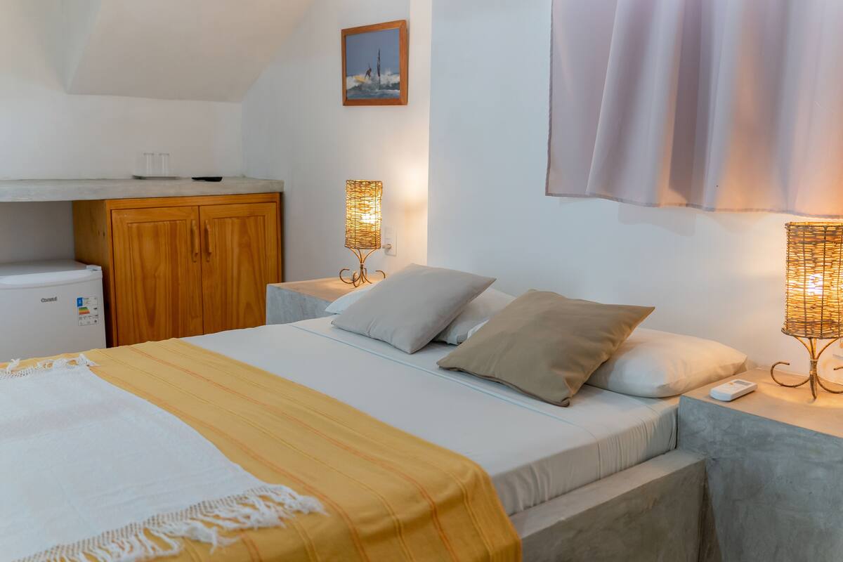 Quarto com cama, mesas de cabeceira, armário, luminária e uma pequena cozinha compacta.