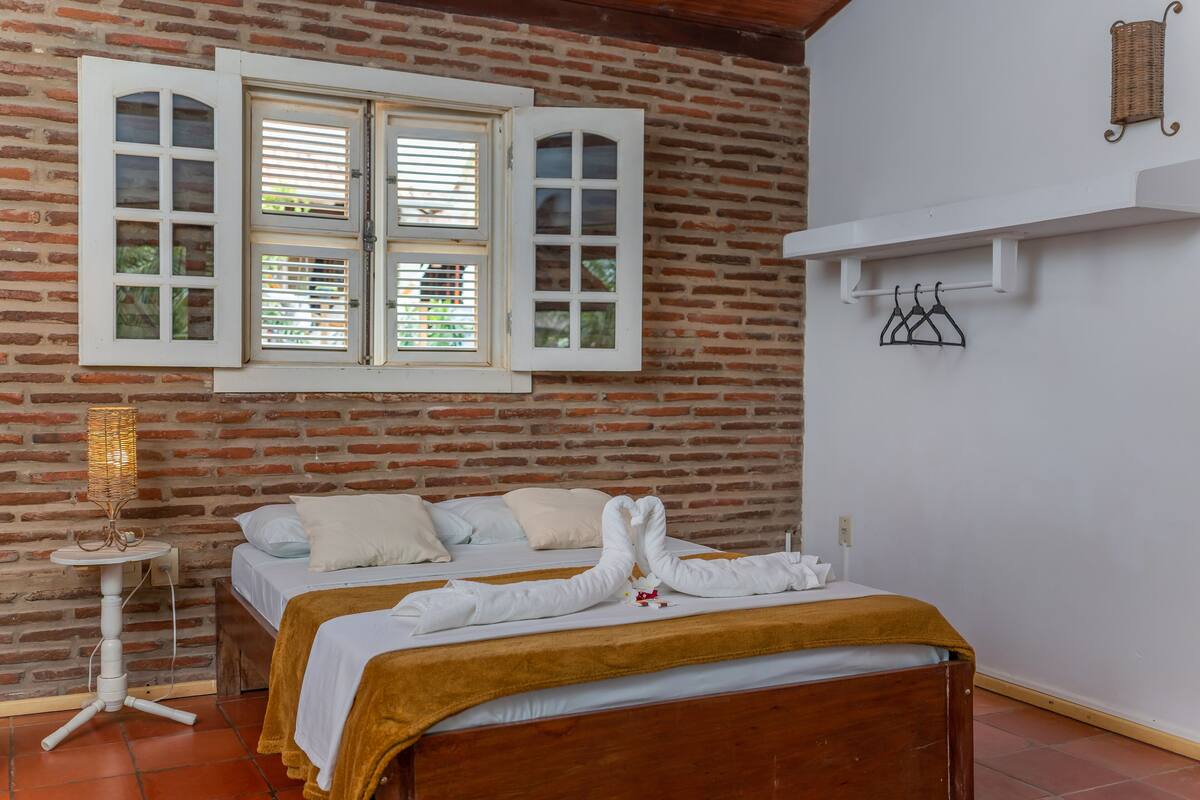 Quarto de hotel com duas camas, parede de tijolos, janela com persianas, pequena mesa com abajur e prateleira com cabides.