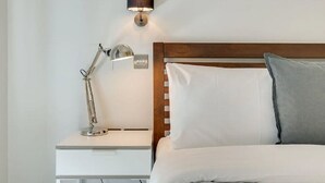 Una cama bien hecha con almohadas blancas y grises, cabecera de madera, lámpara de noche y aplique de pared.