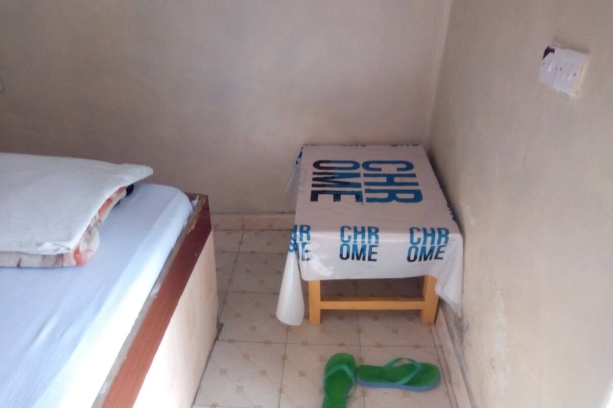 A small room with a bed, a small table with a blanket, and a pair of slippers on the floor.