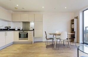A modern kitchen with a dining area, wooden flooring, and a view through a large window.