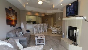 Ein Wohnzimmer mit grauem Sofa, Couchtisch und Kamin. Im Hintergrund befindet sich eine Küche mit weißen Schränken und ein Essbereich mit weißen Stühlen.