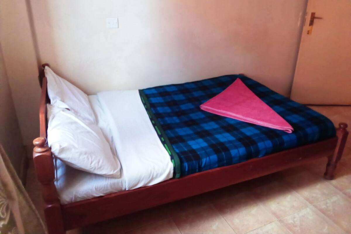 Una cama individual con estructura de madera, una colcha azul y negra a cuadros, una almohada blanca y una toalla rosa doblada.