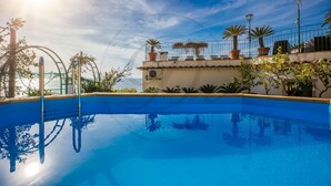 A clear blue pool with a view of the sea, surrounded by potted plants and a building with a tiled roof.