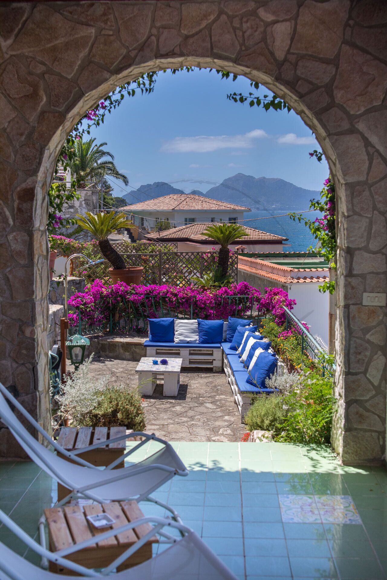 Un arc en pierre mène à une terrasse où se trouvent un canapé bleu, une petite table et une chaise.