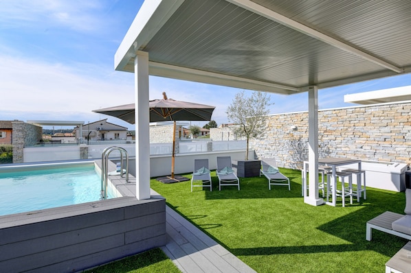 A modern outdoor pool area with a stone wall, a covered seating area with chairs and tables, and a clear view of the surrounding neighborhood.