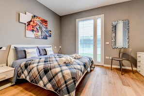 A bedroom with a bed, a chair, a dresser, a window with blinds, and a wall-mounted mirror.