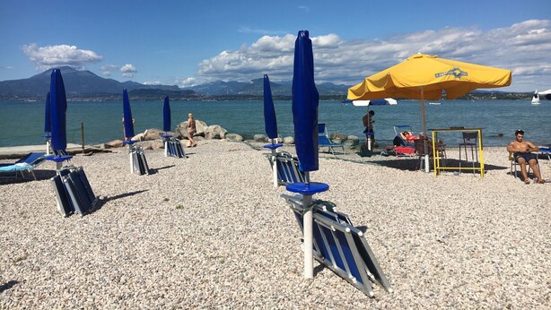 A pebble beach with blue and yellow umbrellas, lounge chairs, and a clear view of the sea and mountains in the background. - Appartamento Rossella (Peschiera del Garda)
