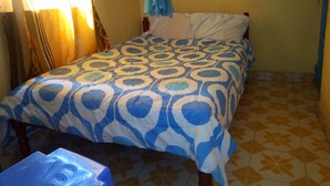 Quarto pequeno com uma cama de solteiro, colcha estampada, cortina e um objeto pendurado na cor azul.