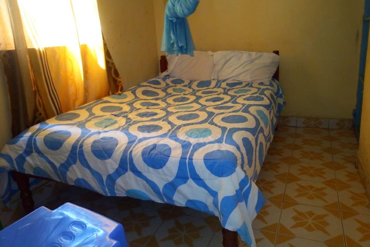 A small, single-bed room with a patterned bedspread, a curtain, and a blue hanging object.