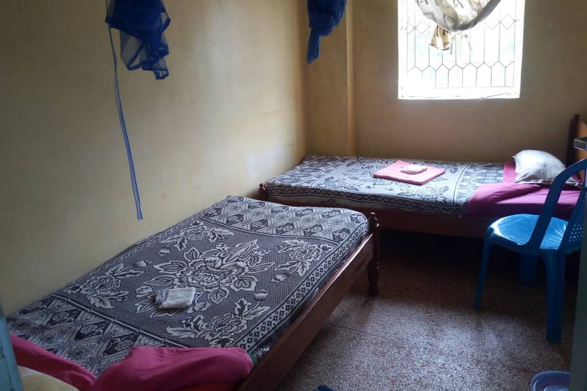 A small room with two single beds, a window with a metal grill, a blue plastic chair, and a curtain.