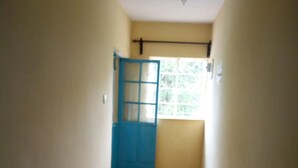 Um corredor estreito com uma porta azul no final, uma janela à direita e chão de azulejos.