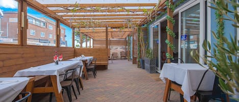 Un patio al aire libre con mesas y sillas de madera, cubierto por una pérgola de madera con luces colgantes.