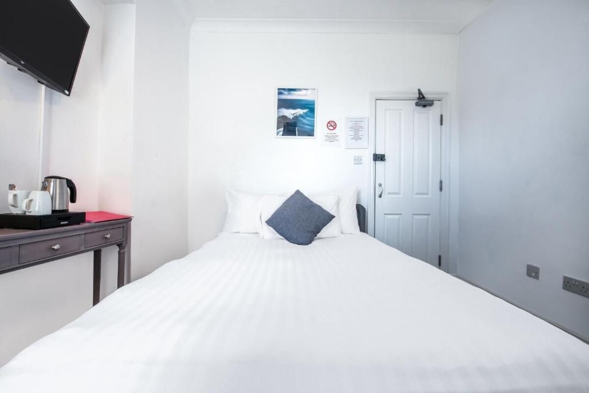 A hotel room with a bed, a desk with a kettle, a flat-screen TV, and a door.