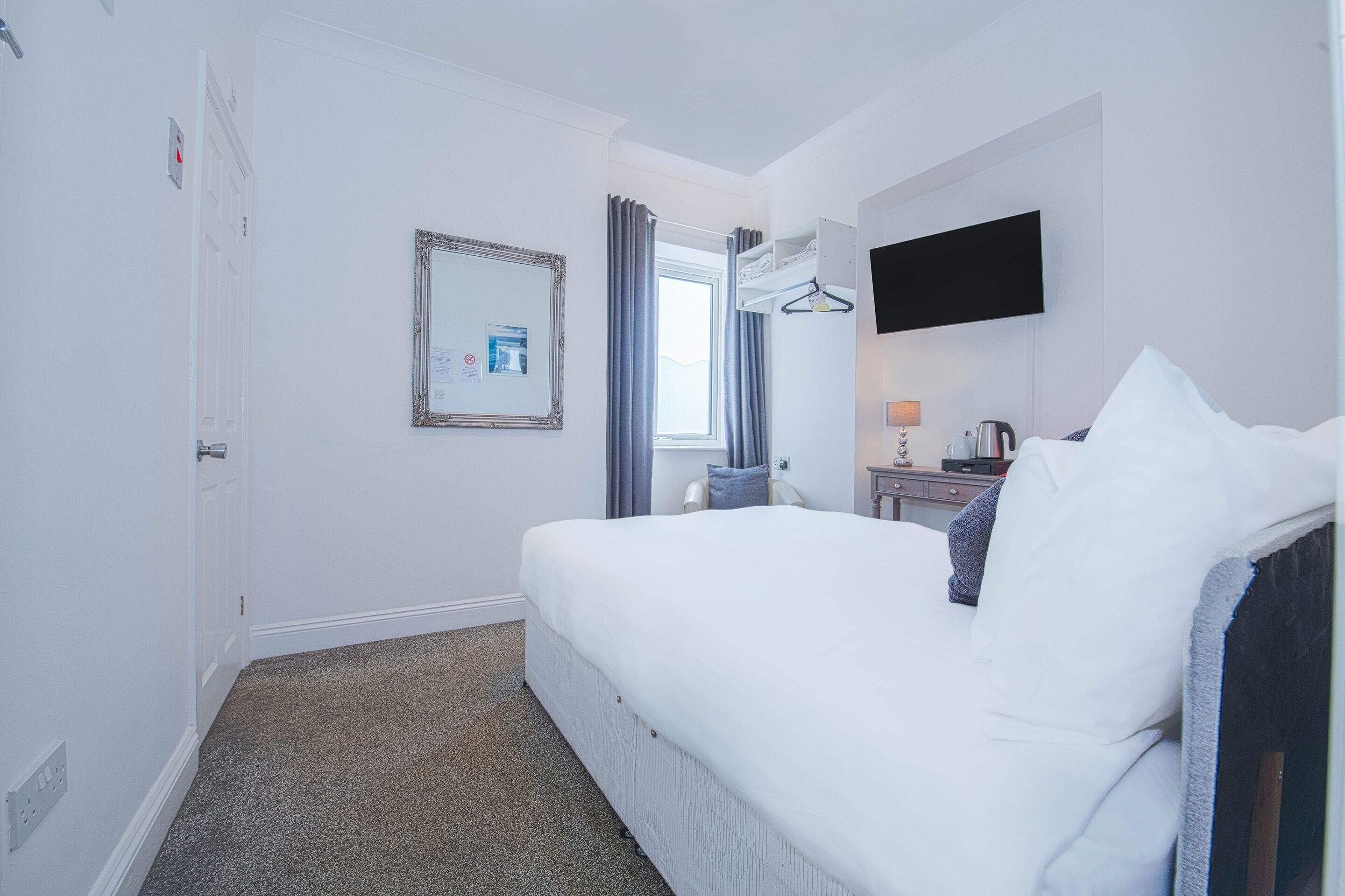 A hotel room with a bed, a flat-screen TV mounted on the wall, a bedside table with a lamp and a coffee maker, a window with curtains, and a door.