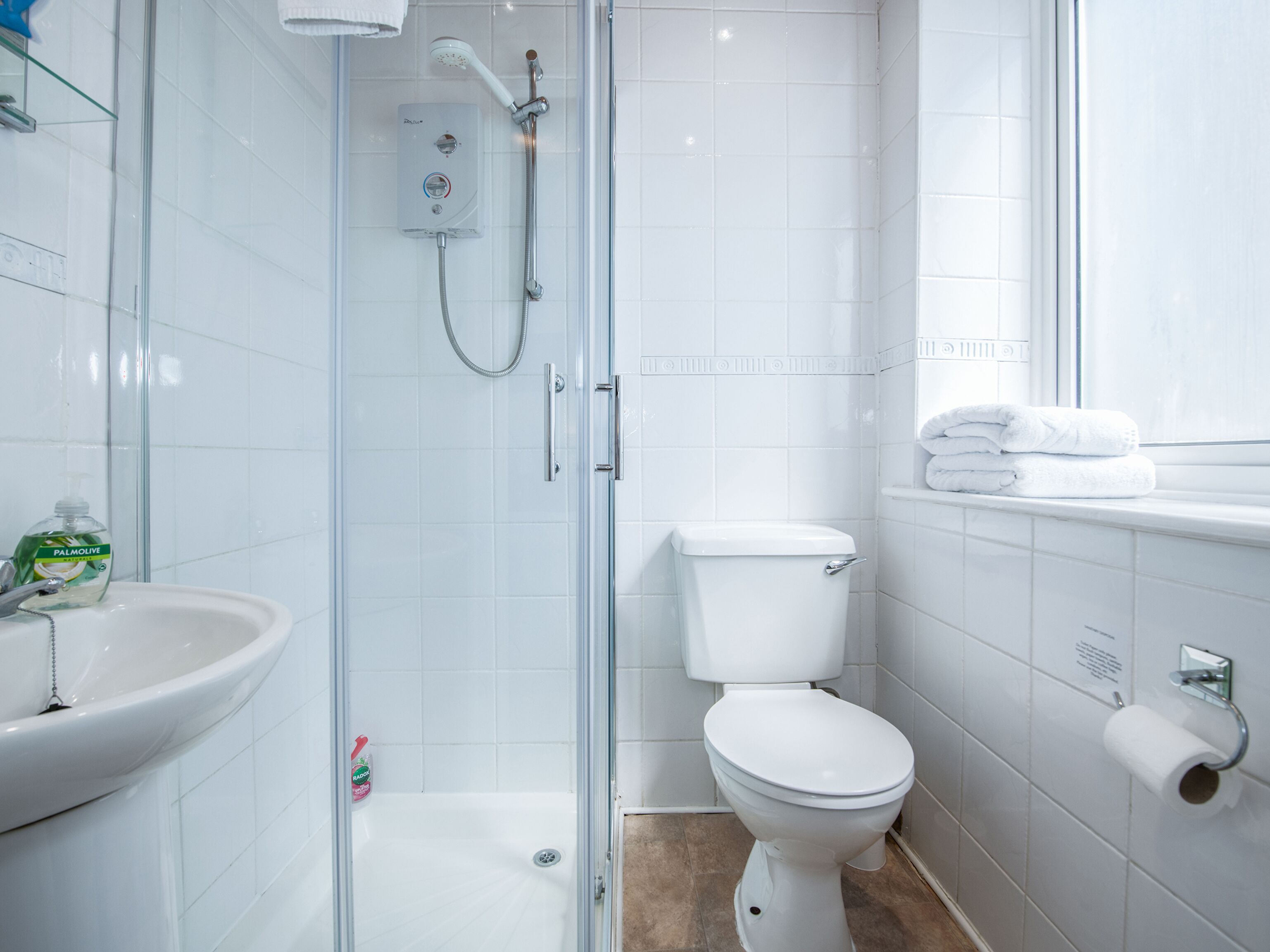 Une salle de bain moderne avec une douche à parois de verre, des toilettes blanches, un lavabo et une fenêtre sur le rebord de laquelle sont posées des serviettes.