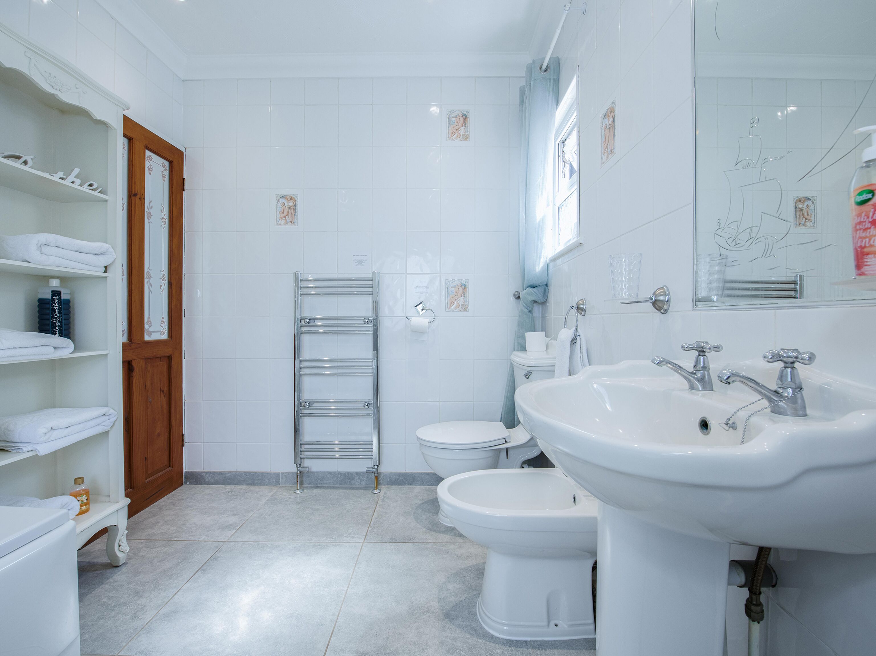A modern bathroom with a toilet, bidet, and a large sink. There are shelves with towels and toiletries.