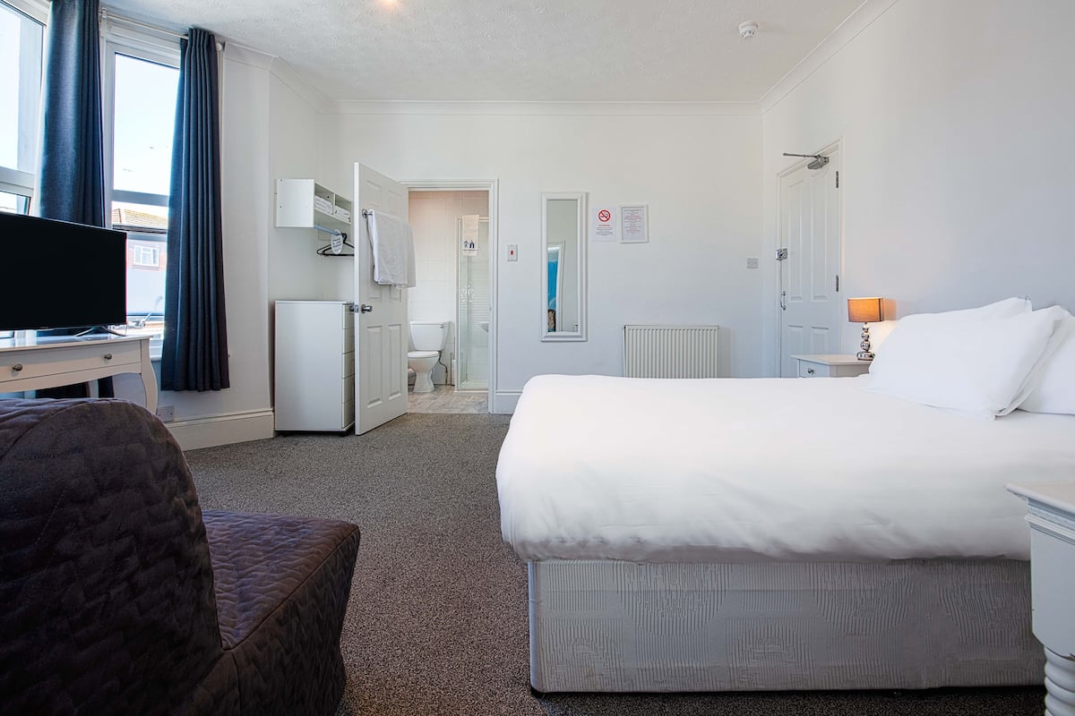 Habitación de hotel con cama, escritorio con televisor, silla, baño con inodoro y ducha, y radiador.