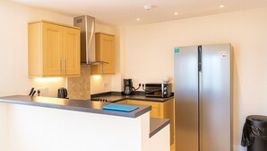 A modern kitchen with wooden cabinets, a stainless steel refrigerator, a coffee maker, and a toaster. - Westbeach Holiday Apartments (Bideford)