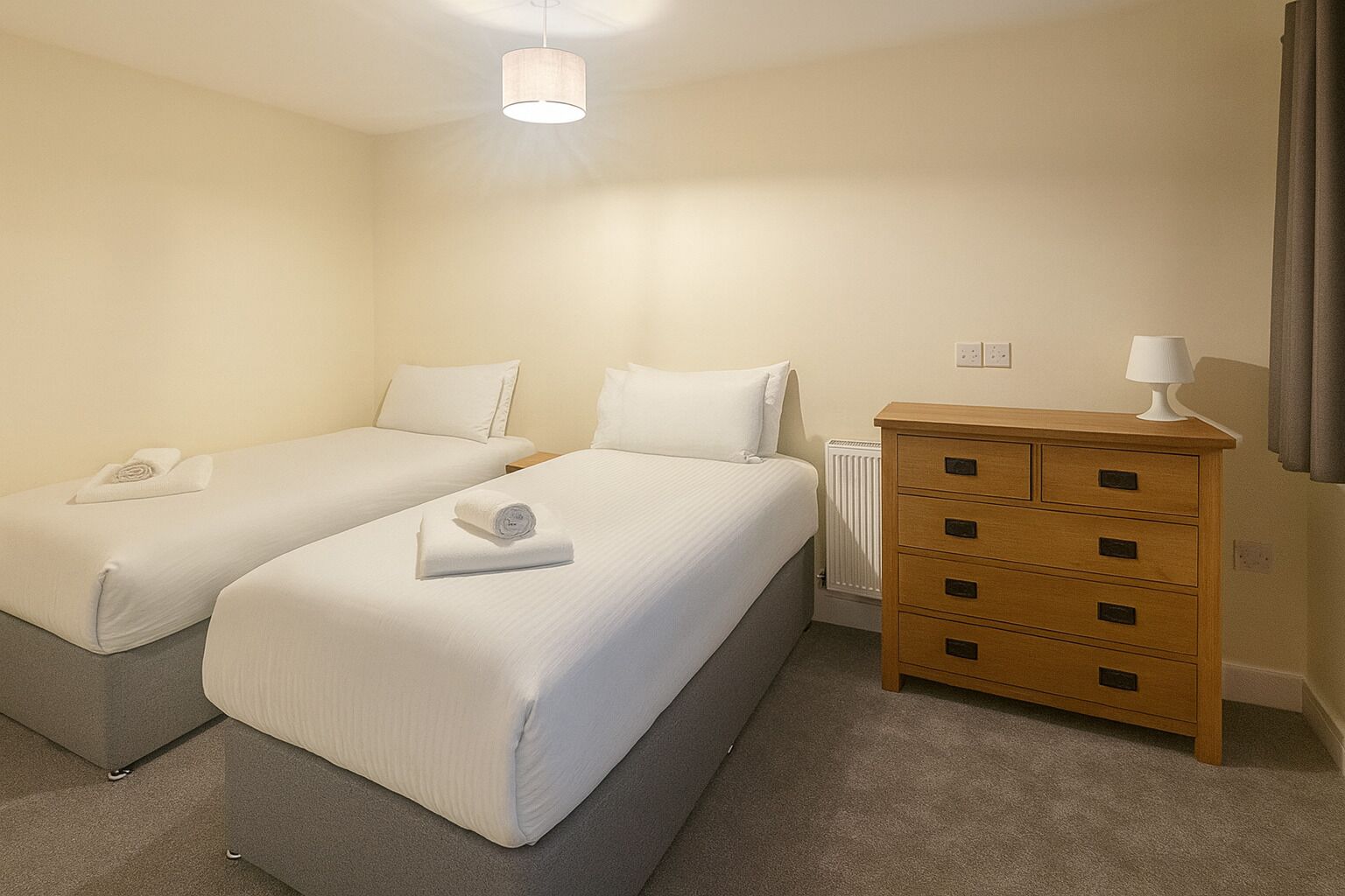 A hotel room with two single beds, a wooden dresser, a radiator, and a lamp.