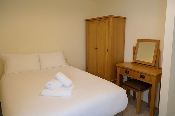A neatly made bed with rolled towels, a wooden wardrobe, and a small wooden desk with a mirror. - Westbeach Holiday Apartments (Bideford)