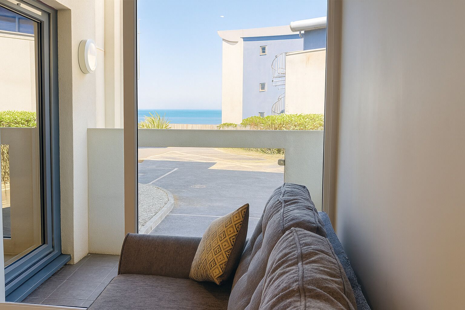 A view from a balcony with a sofa and cushions, overlooking a parking area and a building with a blue facade.