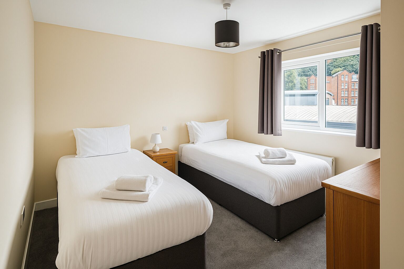 A hotel room with two single beds, a nightstand with a lamp, a wooden desk, and a window with curtains.