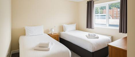 A hotel room with two single beds, a nightstand with a lamp, a wooden desk, and a window with curtains.