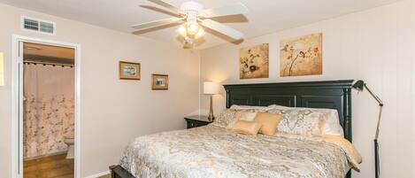 A bedroom with a bed, a ceiling fan, a nightstand with a lamp, and two wall-mounted paintings.