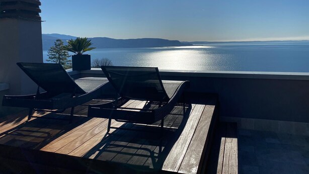 Une terrasse avec deux fauteuils et un pot de fleurs, d’où l’on peut admirer une mer calme et des montagnes lointaines.