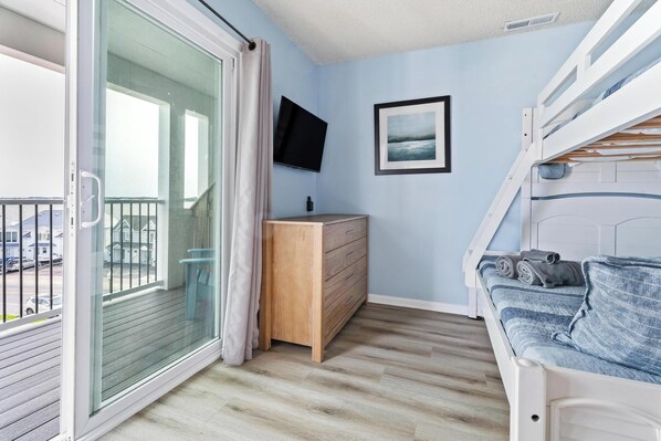 Un balcón con puerta de vidrio corrediza, un aparador de madera, una cama con colchón azul, un televisor montado y un cuadro enmarcado en la pared.