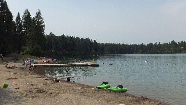 Ein Strand am Seeufer mit Kajaks, einer Anlegestelle und badenden Personen.