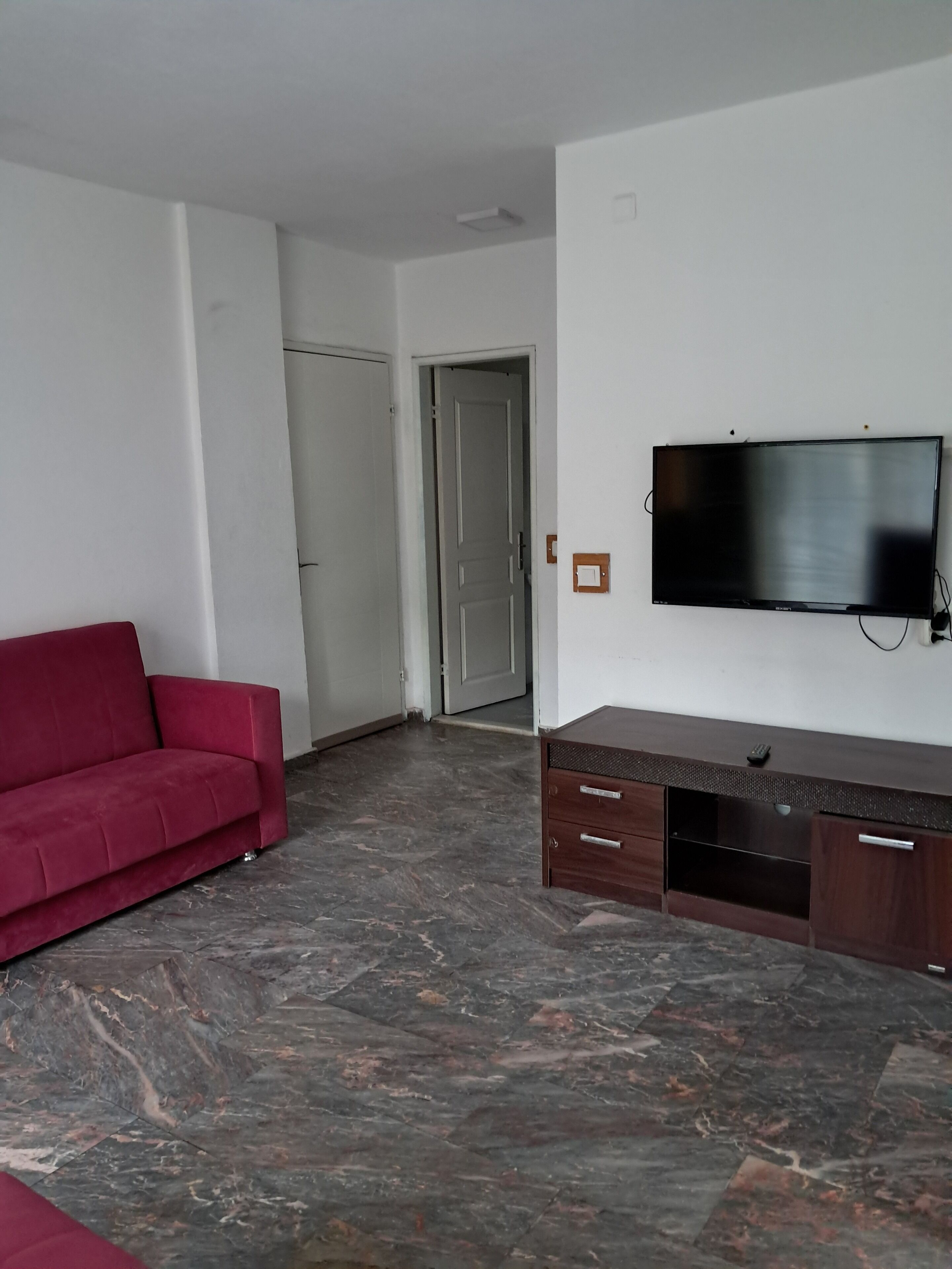 Una sala de estar con un sofá rojo, un televisor de pantalla plana montado en la pared, un mueble de televisión de madera y una puerta que da a otra habitación.