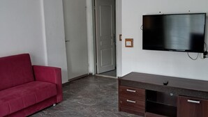 A living room with a red sofa, a flat-screen TV mounted on the wall, a wooden TV stand, and a door leading to another room. - Miramonte Apart Hotel (Marmaris)