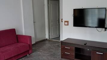 A living room with a red sofa, a flat-screen TV mounted on the wall, a wooden TV stand, and a door leading to another room.