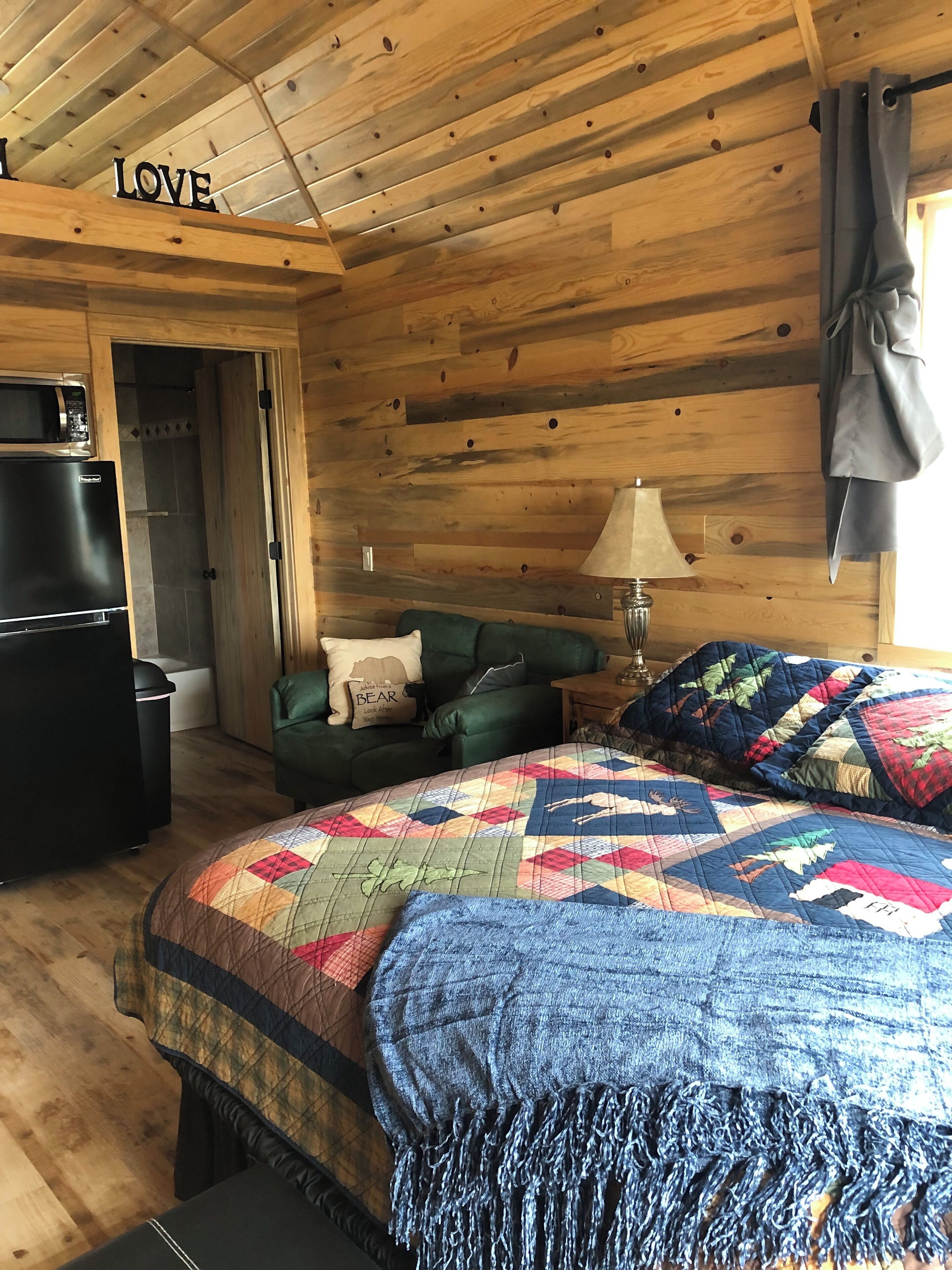 Ein gemütliches Blockhaus-Zimmer mit Holzbalkendecke, einem Bett mit farbenfroher Steppdecke, einem grünen Sofa, einer Lampe und einer Mikrowelle auf einer Arbeitsfläche.