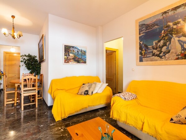 Un salon avec un canapé jaune, une table à manger en bois entourée de chaises, un lustre et un tableau accroché au mur.