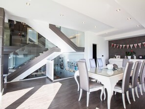 A modern dining area with a glass table and white chairs, a staircase with glass railings, and a kitchen area with holiday decorations.