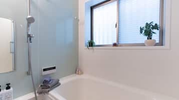 A modern bathroom with a bathtub, a shower, a window with blinds, and a potted plant.