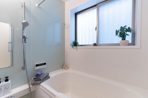 A modern bathroom with a bathtub, a shower, a window with blinds, and a potted plant. - And Koenji (Tokyo)