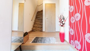 Un couloir avec un escalier en bois, une porte en bois, un miroir, un vase de fleurs et un mur à motifs rouges.