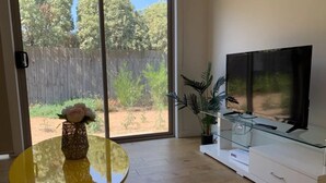 A modern living room with a glass coffee table, a flat-screen TV on a white cabinet, and a sliding glass door leading to a backyard with a wooden fence and greenery. - Delicate Bundoora townhouse 10 (Bundoora)