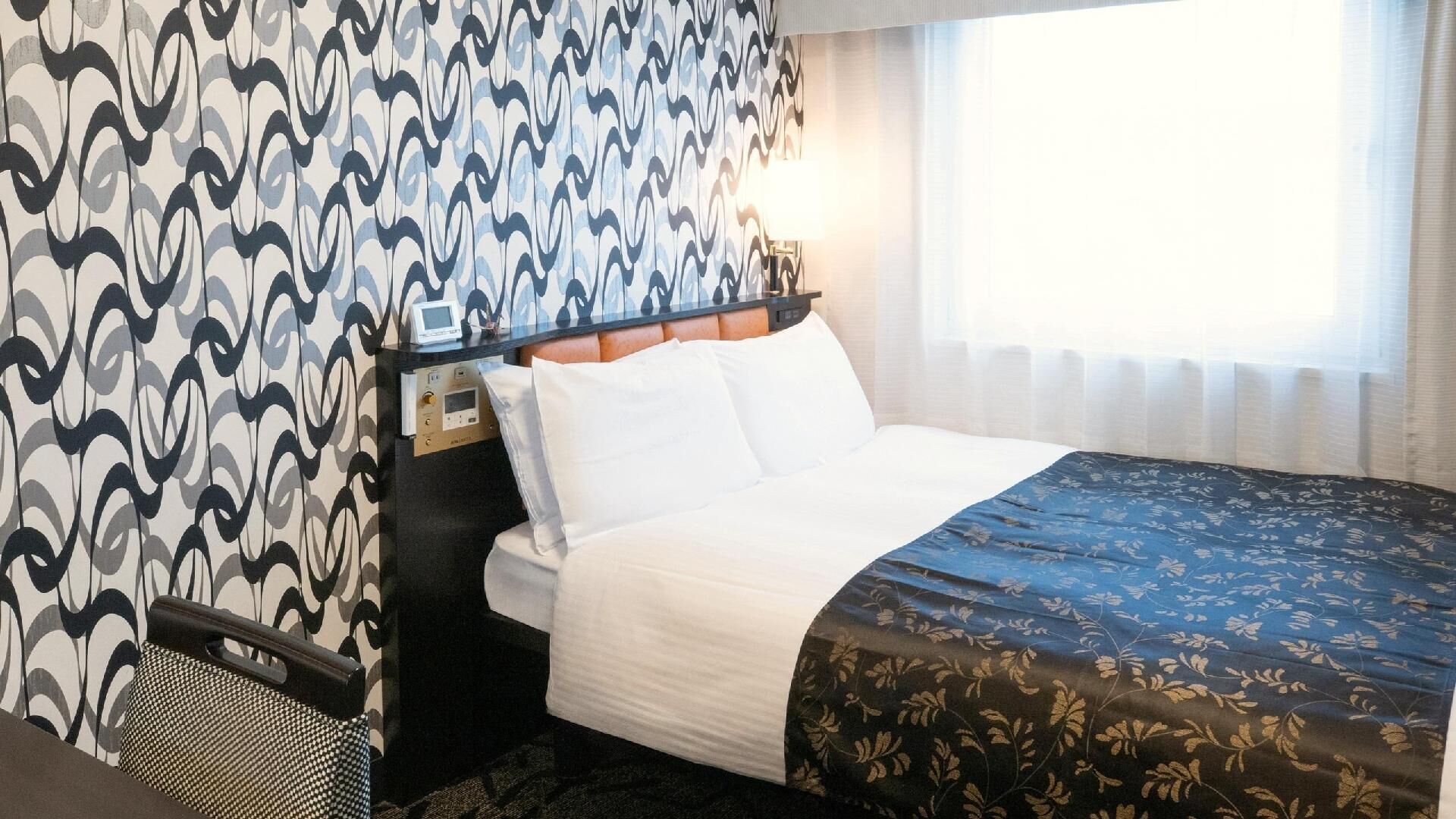 Ett hotellrum med en säng, ett nattduksbord med en kontrollpanel, en lampa och ett fönster med gardiner.