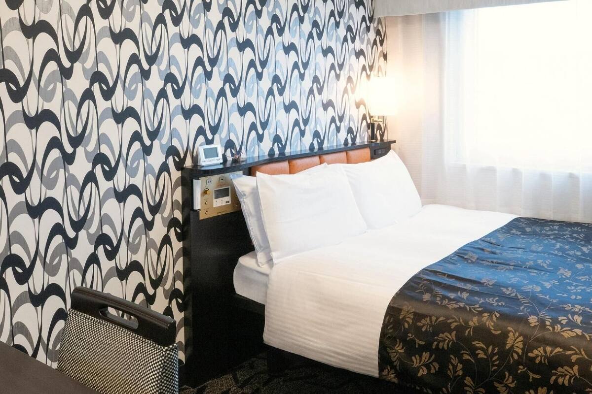 A hotel room with a bed, a bedside table with a control panel, a lamp, and a window with curtains.