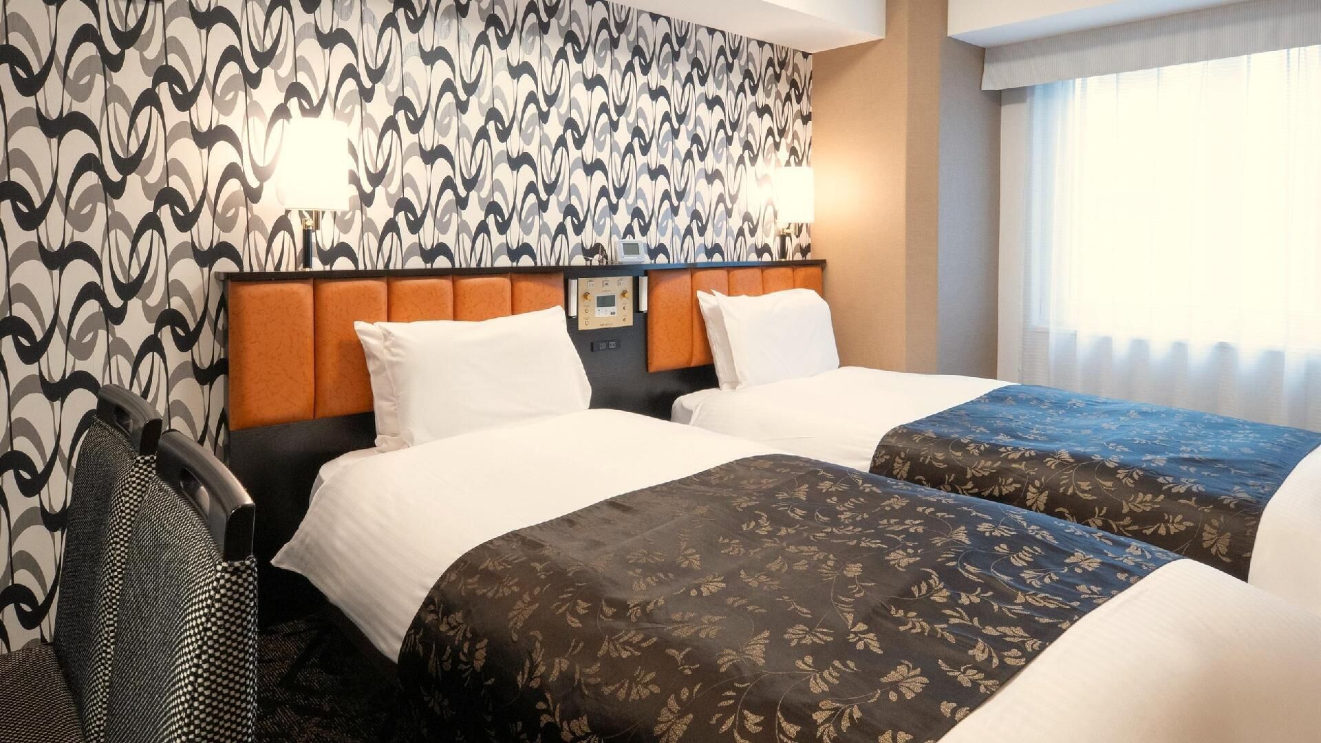 Ett hotellrum med två sängar, en sänggavel med en kontrollpanel, en stol och ett fönster med gardiner.