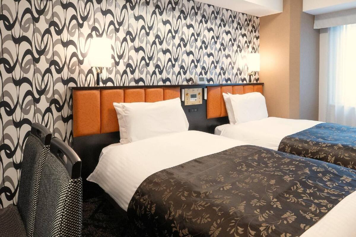 A hotel room with two beds, a headboard with a control panel, a chair, and a window with curtains.